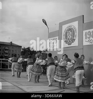 Slawjansk, Ukraine, UdSSR - September 1985: Archivfoto einer Volkstanz-Aufführung während der Stadtfeier. Ein gemischtes Ensemble in traditionellen Trachten tanzt auf der Bühne vor einem Hintergrund mit dem sowjetischen Stadtwappen und Datumsangaben „1676 - 1985“. Auf dem Banner wird ausdrücklich der „8. September“ erwähnt. Das Schwarz-weiß-Bild fängt die festliche Atmosphäre, die dynamische Bewegung der Tänzer und die urbane Landschaft mit einem Bus in der Region Donezk während der friedlichen Ära ein Stockfoto