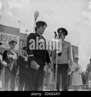 Slawjansk, Ukraine, UdSSR - 8. September 1985: Archivfoto eines jungen Piloten der Zivilluftfahrt, der eine Rede am Mikrofon während des Stadttags hält. Er steht auf der Hauptbühne neben einem Kadett in einem hellen Hemd. Veteranen mit Medaillen und Blumen sind im Hintergrund neben einem Banner mit „1985“ zu sehen. Dieses Schwarz-weiß-Bild fängt das Prestige des Luftfahrtberufs und die feierliche Atmosphäre des Urlaubs in der Region Donezk während der friedlichen Sowjetzeit ein. Stockfoto