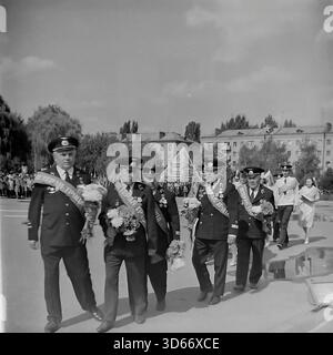 Slawjansk, Ukraine, UdSSR - 8. September 1985: Archivfoto einer festlichen Prozession während des Stadttages. Eine Gruppe von Zivilluftfahrtbeamten mit zeremoniellen Schärpen mit der Aufschrift „Veteran of Labor“ geht über den zentralen Platz mit Blumen. Hinter ihnen sind Wohngebäude und eine Menschenmenge zu sehen. Dieses Schwarz-weiß-Bild fängt den Respekt für die beruflichen Verdienste und die Atmosphäre des Urlaubs in der Region Donezk während der friedlichen Sowjetzeit ein. Stockfoto