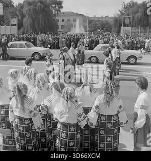 Slawjansk, Ukraine, UdSSR - 8. September 1985: Historisches Archivfoto des Stadtfestes auf dem zentralen Platz. Ein Volkstanz-Ensemble in traditionellen ukrainischen Kostümen begrüßt das Brautpaar, das in einem dekorierten Wolga-Auto ankommt. Das Schwarzweiß-Bild zeigt eine feierliche Zeremonie mit einer Braut und einem Bräutigam während einer öffentlichen Großveranstaltung. Im Hintergrund sind eine große Menschenmenge und ein Brunnen zu sehen, die die kulturellen Traditionen der Donezker Region in der Sowjetzeit widerspiegeln. Stockfoto