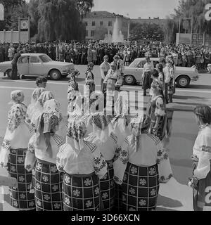 Slawjansk, Ukraine, UdSSR - 8. September 1985: Historisches Archivfoto des Stadtfestes auf dem zentralen Platz. Ein Volkstanz-Ensemble in traditionellen ukrainischen Kostümen begrüßt das Brautpaar, das in einem dekorierten Wolga-Auto ankommt. Das Schwarzweiß-Bild zeigt eine feierliche Zeremonie mit einer Braut und einem Bräutigam während einer öffentlichen Großveranstaltung. Im Hintergrund sind eine große Menschenmenge und ein Brunnen zu sehen, die die kulturellen Traditionen der Donezker Region in der Sowjetzeit widerspiegeln. Stockfoto