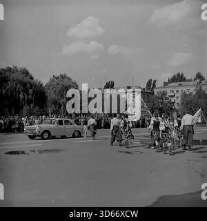 Slawjansk, Ukraine, UdSSR - 8. September 1985: Großes Archivfoto des Stadtfestes auf dem zentralen Platz. Ein weißer GAZ-24 Volga-Wagen fährt an einem Volkstanz-Ensemble in traditionellen Kostümen vorbei, der auf dem Asphalt spaziert. Im Hintergrund steht eine Zuschauermenge in der Nähe einer Propagandapyramide, die dem XXVII. KPdSU-Kongress gewidmet ist. Dieses Schwarzweiß-Bild fängt die urbane Landschaft und die festliche Atmosphäre der Donezker Region während der friedlichen Sowjetzeit ein. Stockfoto
