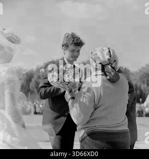 Slawjansk, Ukraine, UdSSR - 8. September 1985: Berührendes Archivfoto eines jungen Bräutigams, der einer älteren Frau während des Stadttags einen Blumenstrauß präsentiert. Links ist ein Fragment des weißen Kleides der Braut zu sehen. Die Szene symbolisiert die Verbindung zwischen Generationen auf dem zentralen Platz. Dieses Schwarz-weiß-Bild fängt einen respektvollen Moment des sowjetischen gesellschaftlichen Lebens und der Traditionen in der Donezker Region während der friedlichen Ära ein. Stockfoto