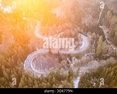 Eine gewundene Straße schlängelt sich durch einen üppigen Wald, während die Sonne untergeht und ein warmes Licht über die Landschaft wirft. Bäume säumen die Route und bereichern die natürliche Schönheit der Gegend. Stockfoto