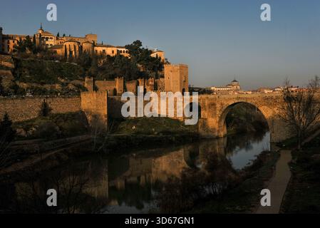 Warme Reflexionen im ruhigen Fluss Tejo von alten Mauerwerken der wiederaufgebauten römischen Puente de Alcántara oder der Alcántara-Brücke, einst der Eingang der Pilger zum UNESCO-Weltkulturerbe Toledo in Kastilien-La Mancha, Zentralspanien. Die Brücke wurde für das römische Toletum erbaut, wurde dann von den Westgoten und Mauren genutzt, mit dem Westtorturm mit königlichen Wappen der spanischen katholischen Monarchen Ferdinand und Isabella aus dem 15. Jahrhundert. Stockfoto
