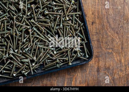 Viele selbstschneidende Schrauben aus goldenem Metall für Bauarbeiten in schwarzem Metalltablett auf hölzernem Hintergrund, Nahaufnahme, Draufsicht, Kopierraum. Neue Befestigungselemente Stockfoto