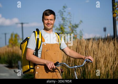 Der lächelnde Kurier in braunem Overall hält einen Fahrradlenker und trägt einen hellgelben Lieferrucksack vor hohen Gräsern und blauem Himmel, was die unabhängige Mobilität in der Stadt symbolisiert Stockfoto