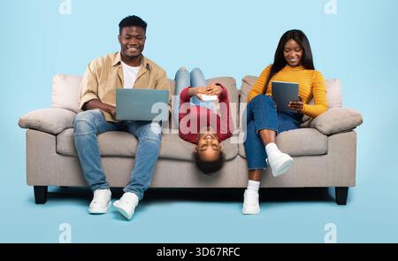 afroamerikanische Familie von drei Leuten, die elektronische Geräte benutzen, auf Sofa auf blauem Hintergrund sitzen Stockfoto