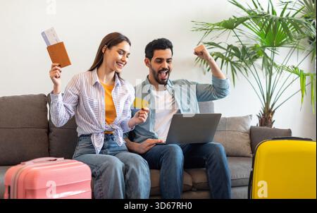 Aufgeregte Touristen paaren sich, die ein Hotelzimmer online buchen, mit Laptop und Kreditkarte, auf dem Sofa mit Koffern sitzen Stockfoto