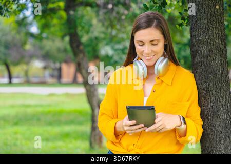 Lächelnde Frau in gelbem Hemd, lehnt sich an einen Baum, liest draußen Tablets, parkt. Stockfoto