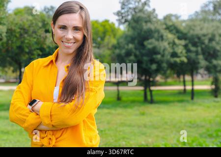 Lächelnde Frau, die draußen im Park mit gekreuzten Armen steht und in die Kamera blickt Stockfoto