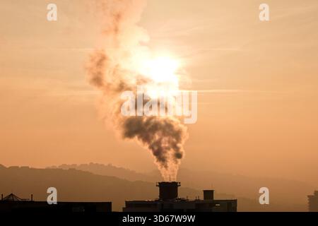 Aus der Vogelperspektive raucht aus einem Industrieschornstein vor dem Hintergrund von trüben Hügeln und einem sonnenverwöhnten Himmel, Sarajevo, Föderation von Bosnien und Herzegowina Stockfoto