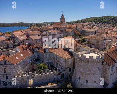 Aus der Vogelperspektive auf sonnengeküsste Steinhäuser mit roten Ziegeldächern, die um eine historische Festung und einen Kirchturm an der Küste gruppiert sind, Korčula, Dubrovnik- Stockfoto