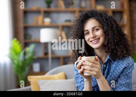 Lächelnde Frau mit lockigen braunen Haaren, die sich auf einem gemütlichen Sofa in ihrem modernen Wohnzimmer entspannt, eine warme Tasse hält und eine friedliche Morgenpause genießt, zufrieden und komfortabel Stockfoto