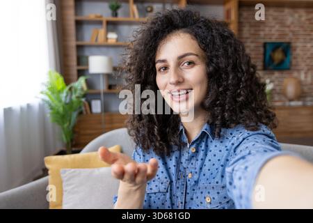 Junge Frau mit lockigem Haar, die sich selbst für ein Video filmt oder an einem Videogespräch teilnimmt, lächelt und mit der Hand Gesten bewegt, während sie von einem komfortablen Wohnzimmer aus kommuniziert Stockfoto