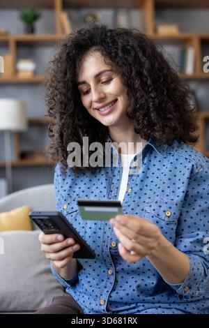 Frau mit dunklen lockigen Haaren lächelt zu Hause, während sie ein Smartphone und eine Kreditkarte hält und eine sichere mobile Zahlung für Online-Shopping mit Leichtigkeit und Selbstvertrauen abschließt Stockfoto