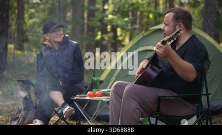 Bärtiger Mann spielt Gitarre für lächelnde Frau, während er im Wald campt, sitzt auf Klappstühlen neben Zelt und Lagerfeuer, mit Tomaten, Gurken und Stockfoto