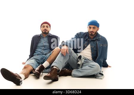 Zwei gutaussehende Brüder posieren selbstbewusst in trendigen Outfits vor reinweißem Hintergrund. Stockfoto
