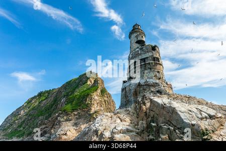 Aus nächster Nähe sehen Sie den verlassenen japanischen Leuchtturm Aniva in Sakhalin, Russland mit rostigen Betonmauern und Seevögeln im felsigen Vordergrund Stockfoto