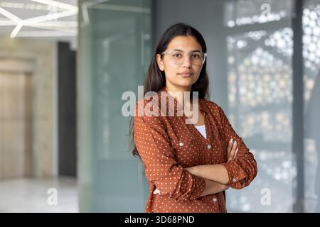 Junge indianerin mit Brille und braunem Hemd, die selbstbewusst mit überkreuzten Armen in einem modernen Bürogebäude steht und Professionalität und Entschlossenheit am Arbeitsplatz repräsentiert Stockfoto