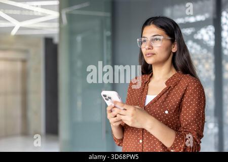 Indianerin mit Brille und braunem Hemd, die ein Smartphone hält und in einem modernen Büro nachdenklich wegschaut, repräsentiert Kommunikation und Technologie im Geschäft Stockfoto