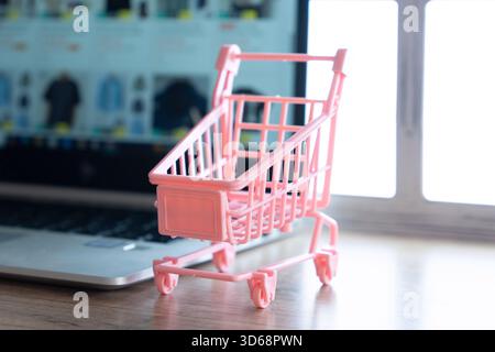 Ein kleiner rosafarbener Einkaufswagen vor einem Laptop mit einer Online-Shopping-Website. Online-Shopping, digitaler Einzelhandel, E-Commerce-Konzept. Stockfoto
