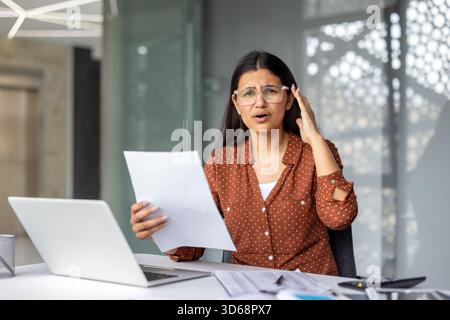 Die junge indische Frau fühlt sich beunruhigt und überwältigt, während sie an ihrem Schreibtisch Papiere überprüft und Ausgaben berechnet, und sieht sich finanziellen Schwierigkeiten und schlechten Nachrichten gegenüber Stockfoto
