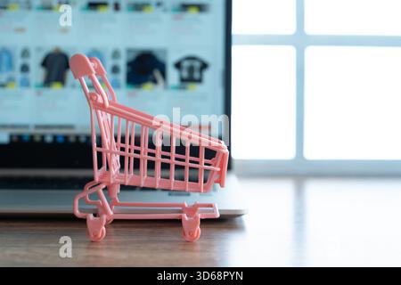 Ein kleiner rosafarbener Einkaufswagen vor einem Laptop mit einer Online-Shopping-Website. Online-Shopping, digitaler Einzelhandel, E-Commerce-Konzept. Stockfoto