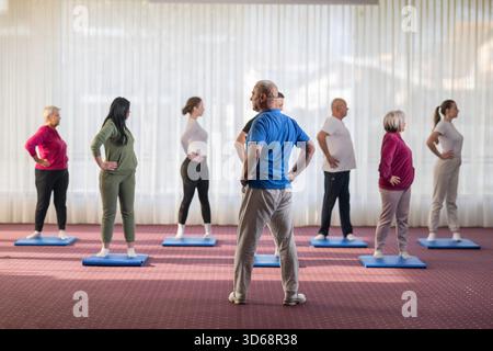 Trainer führt eine vielfältige Gruppe von Menschen während eines Fitnesstrainings mit Dehnungsübungen auf Matten in einem hellen Wellnesscenter. || Modell freigegeben Stockfoto