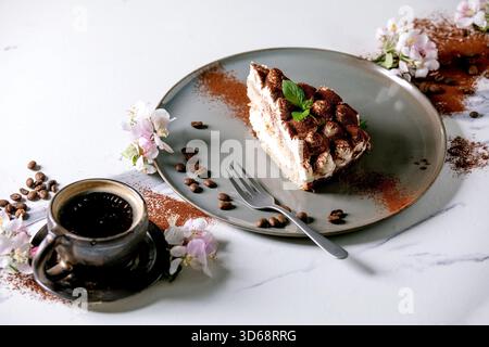 Hausgemachtes, glutenfreies Tiramisu traditionelles italienisches Dessert, bestreut mit Kakaopulver, dekoriert mit blühendem Apfelbaum, Kaffee, Minzblättern und Kaffeebohnen auf weißem Marmorhintergrund. Stockfoto