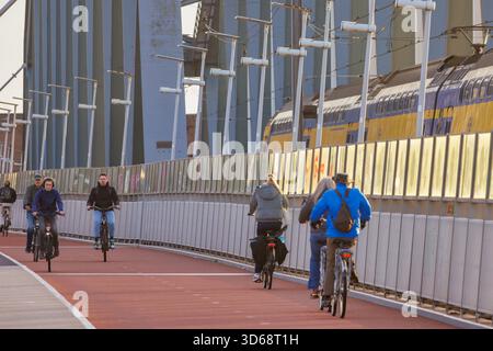 Nijmegen, Niederlande - 12. November 2025: Menschen radeln neben einem vorbeifahrenden Personenzug auf einer Brücke in Nijmegen, Niederlande Stockfoto