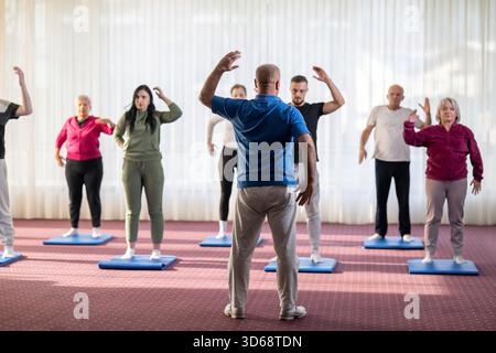 Trainer führt eine vielfältige Gruppe von Menschen während eines Fitnesstrainings mit Dehnungsübungen auf Matten in einem hellen Wellnesscenter. || Modell freigegeben Stockfoto