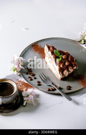 Hausgemachtes, glutenfreies Tiramisu traditionelles italienisches Dessert, bestreut mit Kakaopulver, dekoriert mit blühendem Apfelbaum, Kaffee, Minzblättern und Kaffeebohnen auf weißem Marmorhintergrund. Stockfoto