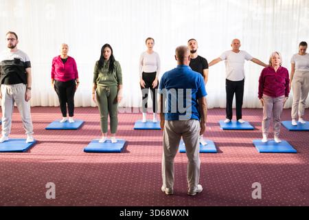 Trainer führt eine vielfältige Gruppe von Menschen während eines Fitnesstrainings mit Dehnungsübungen auf Matten in einem hellen Wellnesscenter. || Modell freigegeben Stockfoto