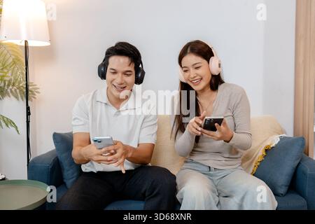 Ein fröhlicher Mann und eine Frau, die schnurlose Kopfhörer tragen, genießen Musik auf ihrem Smartphone, während sie zusammen auf einem gemütlichen Sofa sitzen und lachen Stockfoto