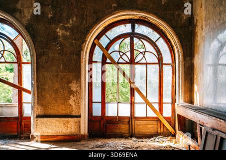 Bogenförmige Holzfenster in verlassenem historischen Gebäude, veraltete Wandstruktur und natürliches Sonnenlicht durch mattiertes Glas, Vintage-Architektur. Stockfoto