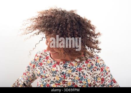 Fröhliche Frau mit lockigem Haar, die während des Lächelns in Bewegung gehalten wird, trägt eine farbenfrohe Bluse mit Blumenmuster vor weißem Hintergrund. Glücksbegriff ener Stockfoto