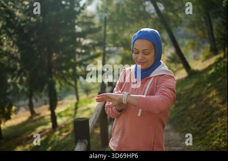 Eine Frau in blauem Hijab und rosafarbenem Hoodie schaut ihre Smartwatch in einem sonnigen Park und veranschaulicht den modernen Alltag einer muslimischen Frau in Europa. Stockfoto