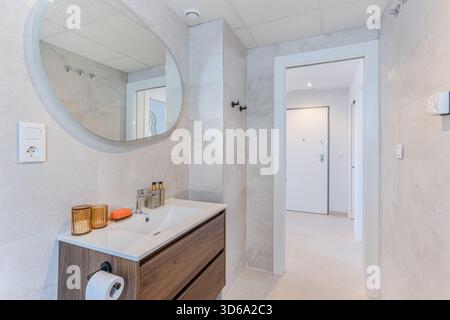 Modernes Bad mit Holzwaschtisch, ovalem Spiegel, dezenter Beleuchtung und minimalistisch gefliesten Wänden Stockfoto