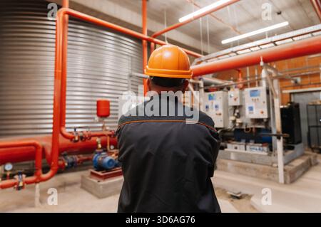 Ingenieur mit Schutzhelm zur Inspektion des industriellen Pumpenraums mit roten Rohren und Schalttafeln Stockfoto