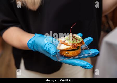 Eine Person, die blaue Handschuhe trägt, präsentiert eine Gourmet-Vorspeise auf einem transparenten Tablett. Das Gericht enthält kunstvoll angeordnete Zutaten, die auf eine Fokussierung hinweisen Stockfoto