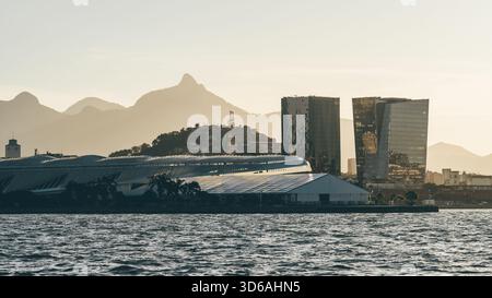 Moderne Skyline am Wasser mit reflektierenden Hochhäusern, futuristischen Dachstrukturen und entfernten Bergsilhouetten, die im warmen Sonnenuntergang leuchten Stockfoto