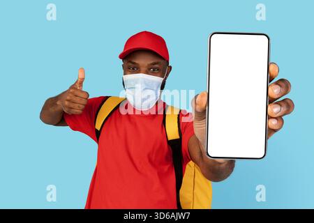Sichere Lebensmittelzustellung. Schwarzer Kurier Mann Trägt Medizinische Maske Zeigt Blank Smartphone Stockfoto