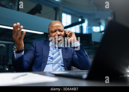 Gestresster afroamerikanischer Geschäftsmann, der am Arbeitsplatz im Büro telefoniert Stockfoto