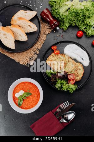 Tomatensuppe und Zucchini-Krapfen mit Joghurtsauce, Salat und Brot auf dunklem Tisch Stockfoto
