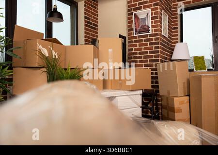 Modernes leeres Wohnzimmer mit Kartonkisten und Möbeln, die in der Nähe der freiliegenden Ziegelwand gestapelt sind. Stapel brauner Kartons, die zum Umziehen und Lagern verwendet werden, angeordnet in leerem Wohnstudio. Stockfoto