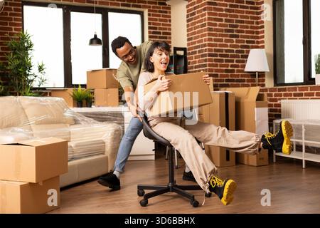 Lächelnde Partner machen Erinnerungen an den bewegten Tag und reiten mit Lachen und spielerischer Energie durch das Wohnzimmer. Sorgloses Paar lacht, als schwarzer Mann Frau auf Stuhl durch leere neue Wohnung schiebt. Stockfoto