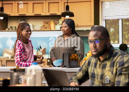 Schwarze Frau und ihre Tochter bereiten Frühstück für Vater und Bruder vor, diskutieren mit der Familie und verbinden während der Morgenroutine. Mutter macht die Kinder fertig mit der Essensvorbereitung. Stockfoto