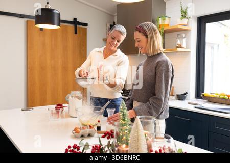 Seniorenmutter und Tochter, die Mehl und Messbecher auf der Kücheninsel absieben Stockfoto