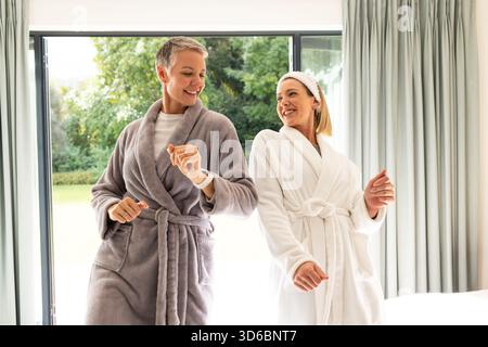 Mutter und Tochter tanzen im Schlafzimmer mit Bademänteln und Smartwatch an Glastür und Vorhängen Stockfoto